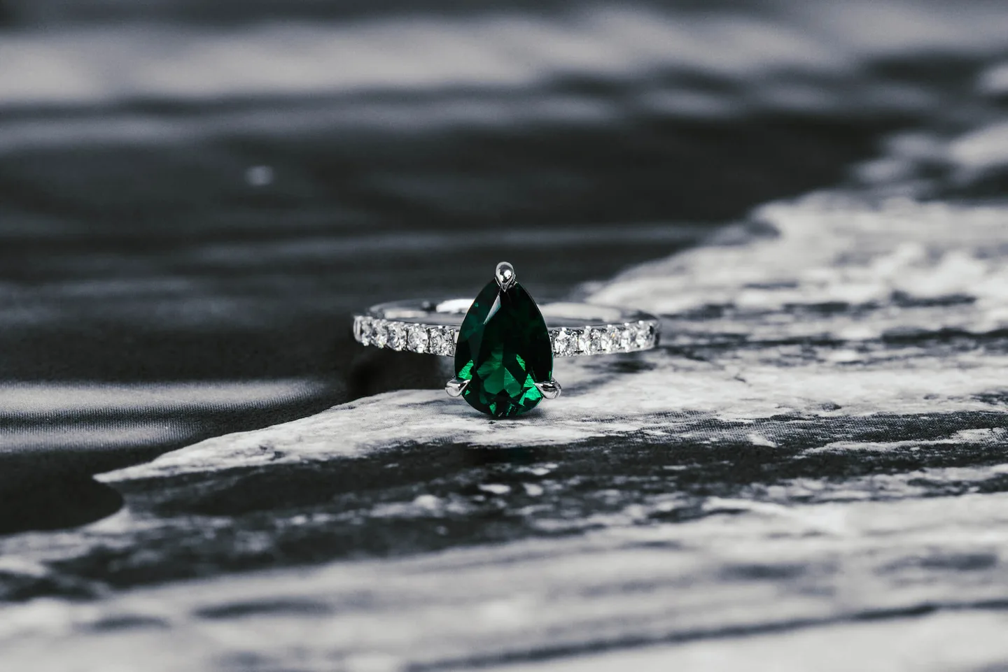 Pear-cut green emerald engagement ring in white metal with a row of round diamonds along the band, on dark marble