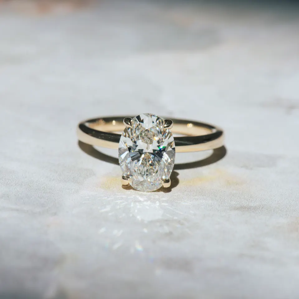 Oval diamond solitaire engagement ring in four yellow gold claw prongs on a slim yellow gold band, on white marble