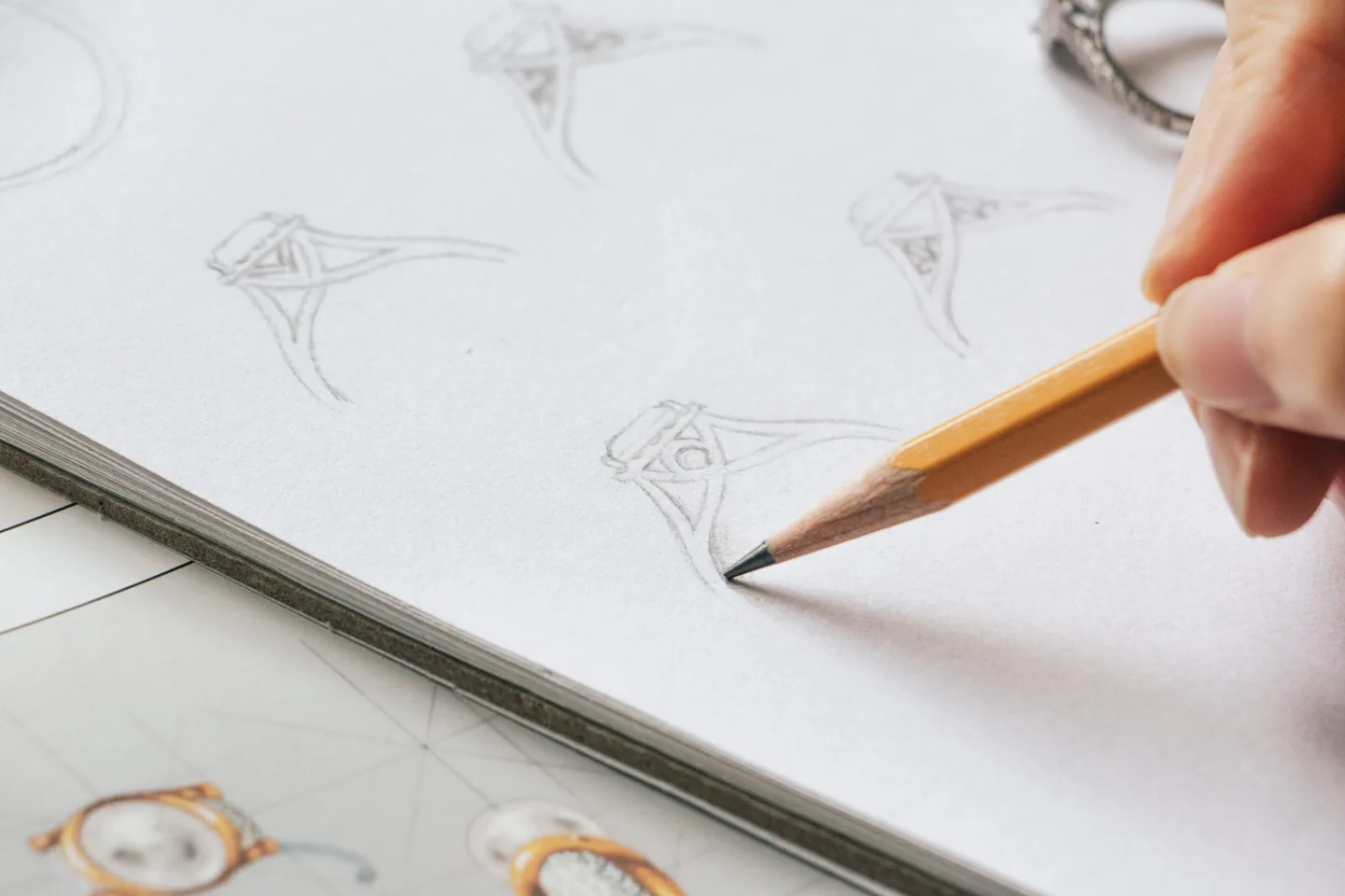 Hand sketching an engagement ring design in pencil beside printed ring photos