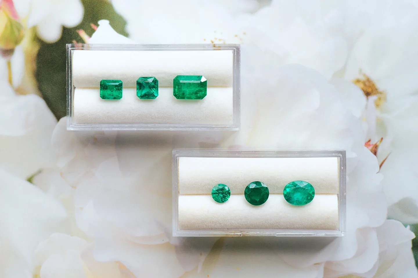 Natural green emeralds, emerald-cut and round, in two white trays beside white flowers