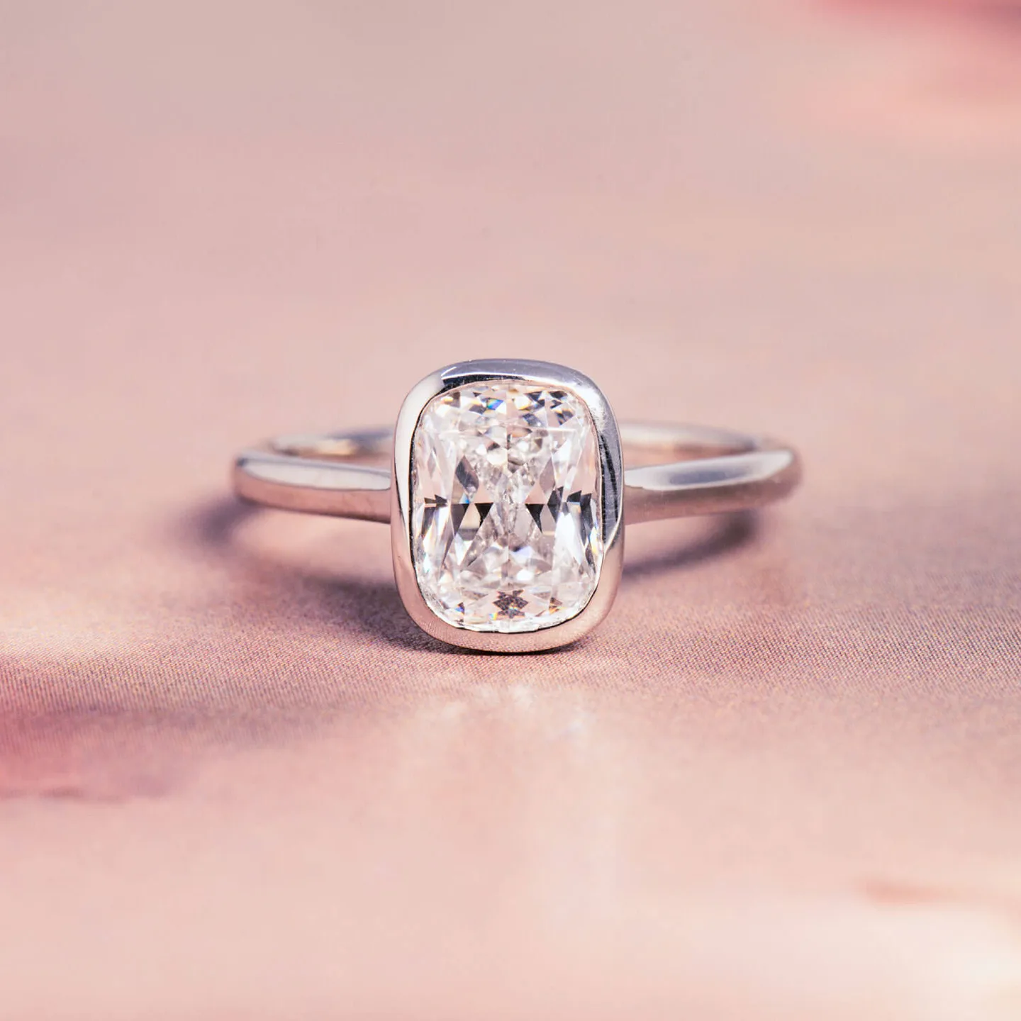 Cushion-cut moissanite bezel-set solitaire engagement ring in white gold on a pink backdrop