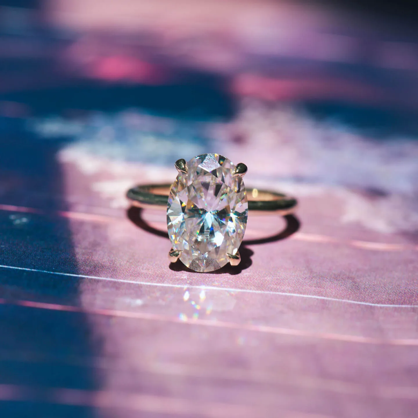 Oval moissanite solitaire engagement ring in yellow gold on a purple and blue backdrop