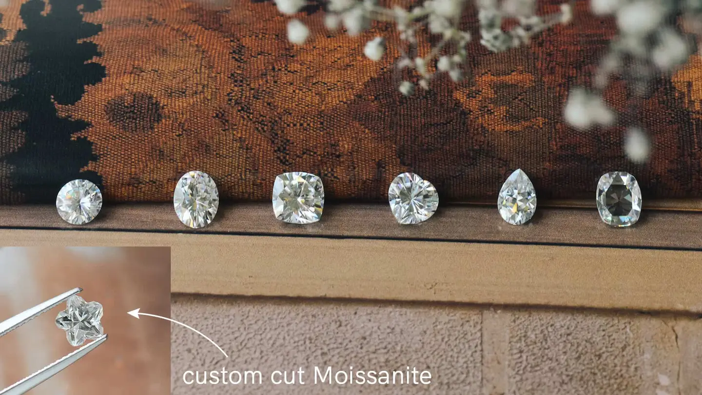 Loose moissanite stones in round, oval, cushion, pear and other cuts on a wooden bench, one custom cut held in tweezers