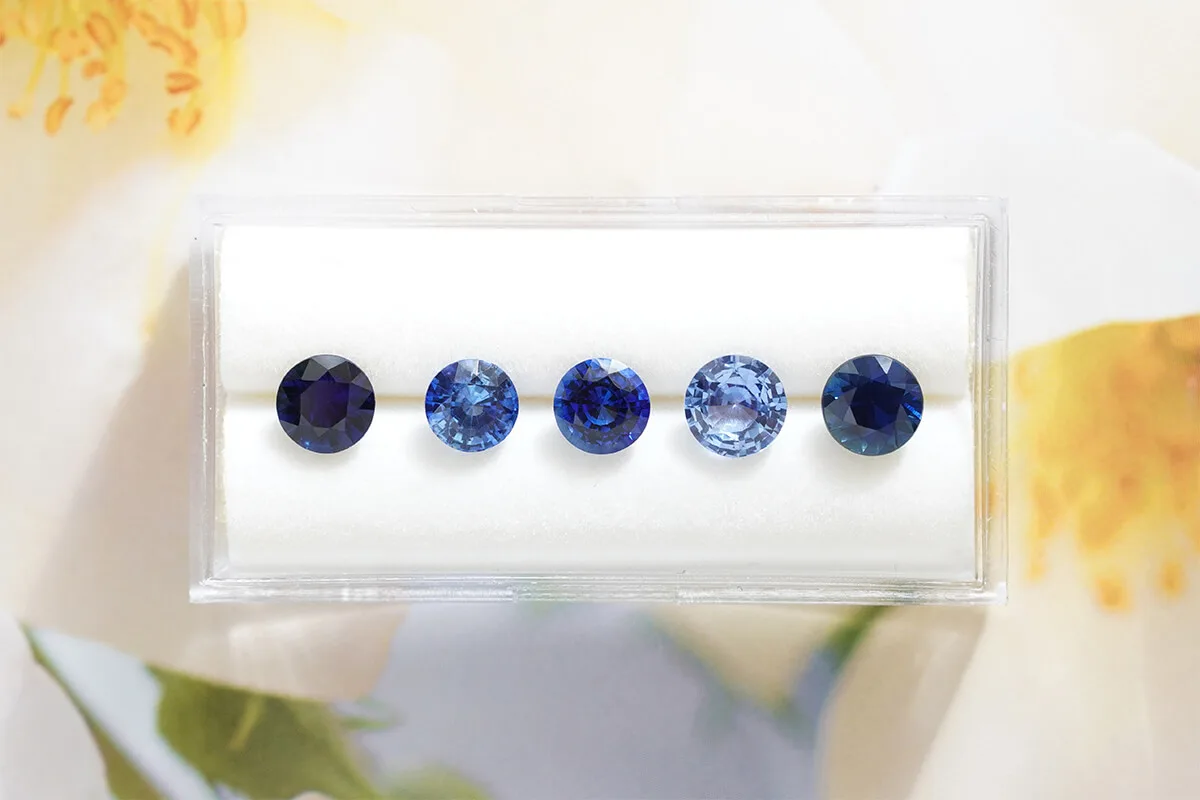 Five round blue sapphires in a white tray, shading from pale to deep blue, beside yellow flowers