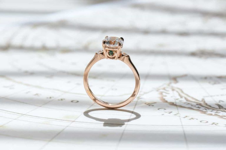 A round-cut rose gold engagement ring featuring a compass logo under the center stone. A tiny rhodolite garnet and emerald are on each side, placed atop a crescent.