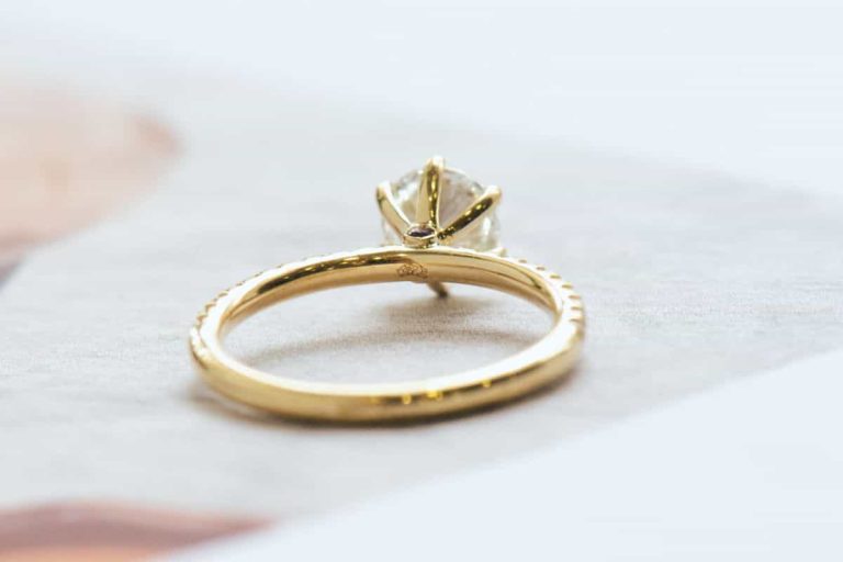 A round-cut rose gold engagement ring featuring a compass logo under the center stone. A tiny rhodolite garnet and emerald are on each side, placed atop a crescent.